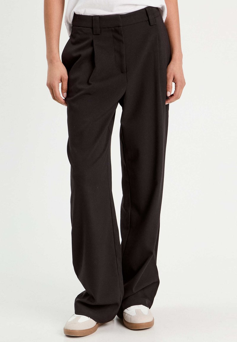 Pantaloni neri a gamba larga realizzati in tessuto liscio, con pieghe frontali, tasche laterali e una cintura su misura. Indossati con scarpe di colore chiaro.