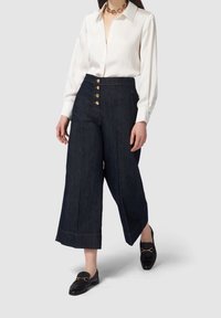 Džínové culottes v tmavě modré barvě, s akcenty zlatých knoflíků, kontrastním šitím a širokým střihem, doplněné bílou košilí s knoflíky.