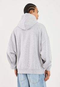 Felpa grigia oversize con cappuccio con cordino. Tessuto morbido e testurizzato. Spalle a caduta e orlo a coste, abbinata a jeans di denim chiaro.