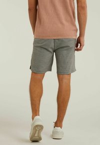 Muž v šedých kostkovaných šortkách, světlém hnědém pleteném topu a bílých teniskách, zobrazen ze zadního pohledu na jednoduchém světlém pozadí.