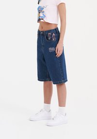 Bermudas de mezclilla en azul oscuro con bordado floral en los bolsillos y costados. Presentan un corte relajado, presillas para cinturón y cierre de botón frontal.