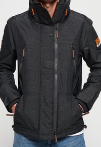 Chaqueta gris oscuro con un alto cuello, frontal con cremallera, dos bolsillos en el pecho con cremallera y acentos naranjas. Tejido texturizado con cordones ajustables.