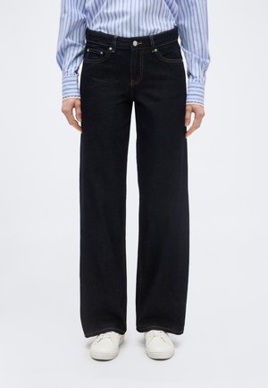 Isik, kes kannab musta laia säärega teksapükse, sinvalget triibulist särk sisse panduna ja valgeid tossusid, seisab tavalise tausta ees.