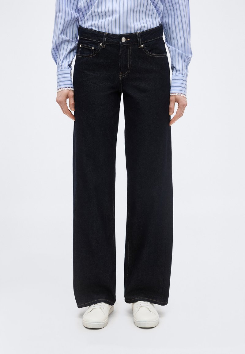 Άτομο που φοράει μαύρα τζιν με φαρδύ πόδι, μπλε και λευκό ριγέ πουκάμισο μπα tucked in, και άσπρα αθλητικά παπούτσια, στέκεται μπροστά σε απλό φόντο.