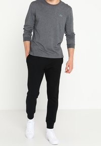 Graues Langarmshirt mit rundem Ausschnitt und grünem Logo, kombiniert mit schwarzen, schmal zulaufenden Jogginghosen und weißen Sneakern.