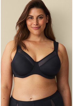 Smilende kvinde med langt hår iført en sort støttende bh og matchende undertøj mod en ensartet beige baggrund.