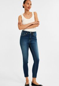 Chude dżinsy w ciemnoniebieskim denimu, o średnim stanie, z pięcioma kieszeniami i kontrastowym przeszyciem; połączone z dopasowanym białym topem na ramiączkach i czarnymi płaskimi butami.