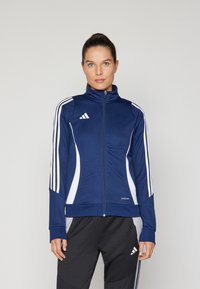 Marinblå idrottsjacka med vita ränder och dragkedja fram. Har hög krage och figurnära design, tillverkad av ett glatt och lätt material.