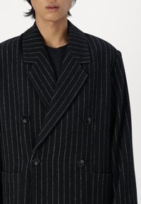 Double-breasted black wool blazer with white pinstripes. Features two front pockets and black buttons. Classic collar design.