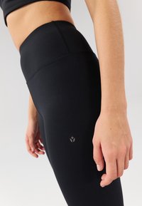 Colanți negri cu talie înaltă, confecționați din material elastic, având o textură fină și un mic logo lângă talie. Vedere de aproape.