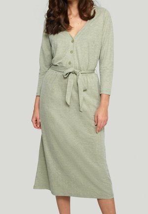 Femme portant une robe midi vert clair à manches longues, boutonnée, avec une ceinture nouée à la taille, sur un fond gris uni.