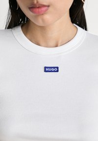 Valkoinen ribattu t-paita, jossa on sininen suorakulmainen "HUGO" -logo-ompelu rinnassa, pyöreä kaula-aukko ja sileä pinta.