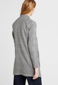 Blazer de cuadro en gris y negro con acentos dorados. Presenta mangas largas, cuello con muesca y corte recto, mostrando un tejido texturizado.