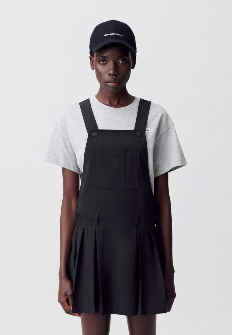 Una joven se encuentra de pie frente a un fondo sencillo, vestida con una camiseta gris y un vestido de tirantes negro, complementado con una gorra negra.