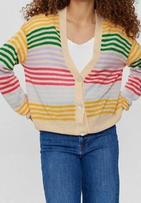 Žena v mnohobarevném pruhovaném kardiganu s knoflíky přes bílý top a modré džíny, s rukama na bocích, vlnité vlasy viditelné.