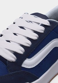 Gros plan sur une basket bleue avec une bande blanche latérale et des lacets blancs serrés sur une tige en tissu texturé.