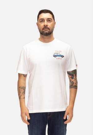 Uomo con capelli corti e scuri e barba che indossa una maglietta bianca con la scritta "Italian Surfer" e una grafica di un'auto blu, in piedi con le braccia ai lati.