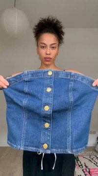 Mlada žena s kovrčavom kosom drži plavu traper suknju s gumbima razvučenu ispred sebe u svijetloj sobi.