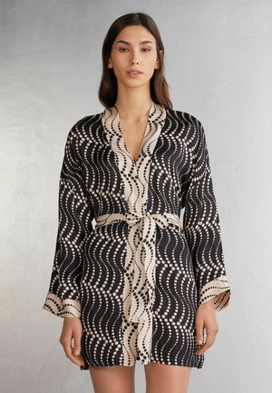Zwart en beige gewatteerde vest met lange mouwen en een strikceintuur, voorzien van een golvende stippenontwerp op een gladde stof.