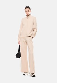 Tuta beige chiaro con giacca con zip e pantaloni a gamba larga, con dettagli testurizzati sulle maniche e sui lati dei pantaloni. Borsa a tracolla nera a forma circolare.
