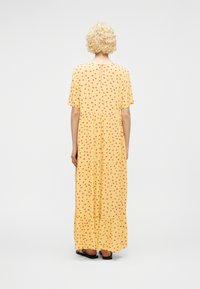 Γυναίκα με κοντά σγουρά ξανθά μαλλιά, ντυμένη με κίτρινο μακρύ φόρεμα με μικρά κόκκινα λουλουδένια σχέδια, που στέκεται γυρισμένη με την πλάτη σε λευκό φόντο.