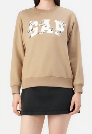Beżowa bluza z napisem "GAP" ozdobionym białymi stokrotkami, zestawiona z czarną mini spódniczką, pokazana na stojącej osobie.