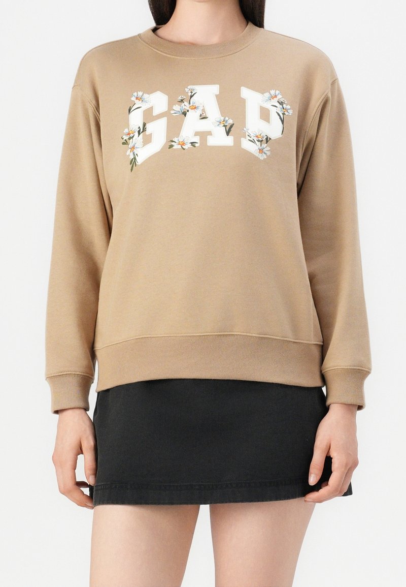 Sweat-shirt beige avec les lettres "GAP" décorées de marguerites blanches, associé à une mini-jupe noire, présenté sur une personne debout.