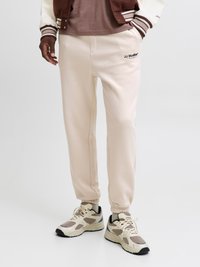 Lichtbeige joggers met elastische boorden en een geborduurd "JJ Studios" logo. Draagt een bruin T-shirt met sneakers in neutrale tinten.
