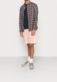 Camisa de franela a cuadros en azul marino y beige sobre una camiseta negra, combinada con pantalones cortos texturizados en rosa claro y zapatillas marrones con detalles en blanco.