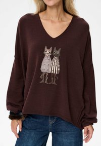 Brązowy sweter z dzianiny z dekoltem w serek, z ilustracją dwóch kotów w wzorzystych sukienkach. Luźny krój, długie rękawy.
