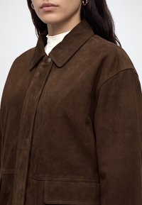 Femme portant une veste en suède marron foncé avec col et fermeture par boutons-pression, un haut blanc à col montant et des boucles d’oreilles créoles dorées.