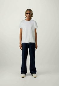 Hvid t-shirt med korte ærmer, parret med løse mørke denimbukser og lyse sneakers. Modellen bærer mørke solbriller. Simple, afslappet design.