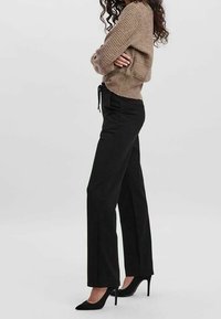 Pantalons noirs à jambes larges avec une texture lisse et une taille à cordon de serrage, associés à un pull en tricot beige et des chaussures à talons hauts noires.