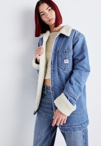 Lichtblauwe denimjas met een witte nepbont kraag en manchetten, voorzien van knoopsluitingen en een borstzak met een label.