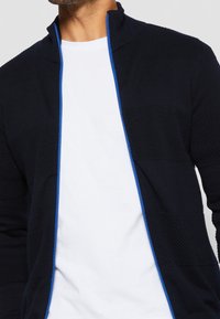 Marinblå stickad zipjacka med texturerade paneler och en kontrasterande blå dragkedja. Bärs över en vit T-shirt, med en hög krage.