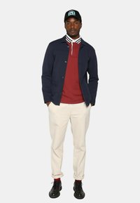 Homme se tenant debout les mains dans les poches, portant une casquette noire, une veste marine, un polo rouge, un pantalon crème, des chaussettes rouges et des chaussures noires.