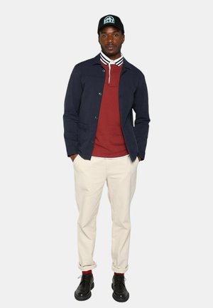 Homme se tenant debout les mains dans les poches, portant une casquette noire, une veste marine, un polo rouge, un pantalon crème, des chaussettes rouges et des chaussures noires.