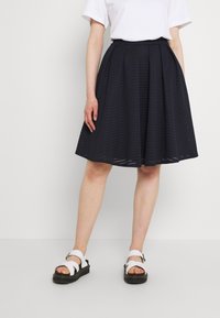 Marinblå veckad kjol i texturerad tyg, med ett vågigt mönster, tillsammans med vita sandaler på en svart sula.