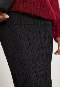 Musta, teksturoitu hame, jossa on ribbineulos. Kangas on aaltoilevakuviollinen ja se on yhdistetty punaisen neulotun neuleen ja mustan clutch-laukun kanssa.