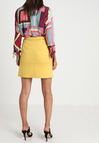 Chemise multicolore à manches longues avec des motifs géométriques, assortie à une jupe jaune. Des escarpins noirs viennent compléter le look.