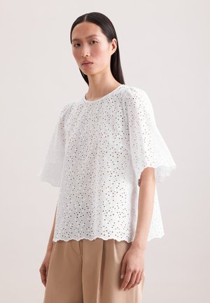 Femme aux cheveux noirs raides portant un chemisier en dentelle ajourée blanc à manches larges et un pantalon beige sur un fond clair.