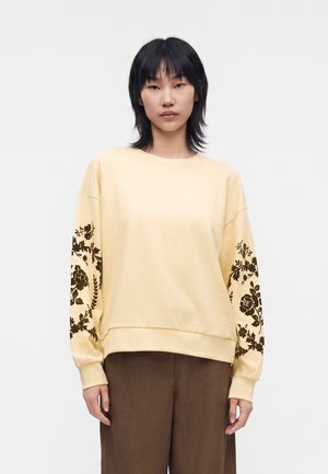 Jeune personne aux cheveux noirs porte un sweat-shirt beige clair avec des motifs floraux noirs sur les manches et un pantalon large marron.
