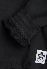 Prenda negra de manga larga con puños doblados que muestran la textura interior y una etiqueta blanca con un panda usando un sombrero de copa.