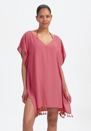 Femme portant un kaftan rose ample avec des franges le long de l'ourlet, debout devant un fond blanc uni.