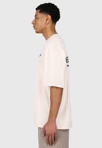 Cremefarbenes, oversize T-Shirt mit kurzen Ärmeln, das ein schlichtes Design und bedruckten Text auf der Rückseite zeigt. Der Stoff wirkt weich und glatt.