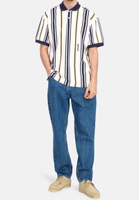 Camisa polo de manga corta con rayas verticales en azul marino, blanco y amarillo, con cuello y un pequeño logo. Se combina con unos vaqueros azules holgados.