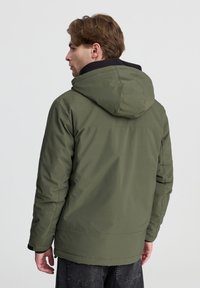 Giacca isolante verde oliva con cappuccio, finitura testurizzata e dettagli neri. Presenta una vestibilità rilassata e colletto leggermente rialzato.