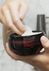 Svart burk med hårbehandling med röd text, hålls i hand. Tjock, vit produkt som skopas ut. Slät, glansig struktur synlig.