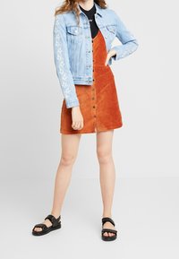 Giacca di jeans blu chiaro con ricami floreali bianchi e un vestito di corduroy arancione con bottoni frontali e una forma svasata. Sandali neri.