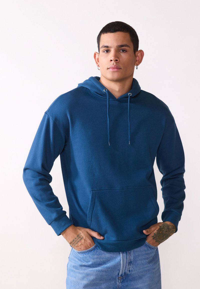 Blauwe katoenen hoodie met een voorzak, een capuchon met koord en geribde manchetten. Draagt met lichtblauwe denim jeans. Soepele textuur.